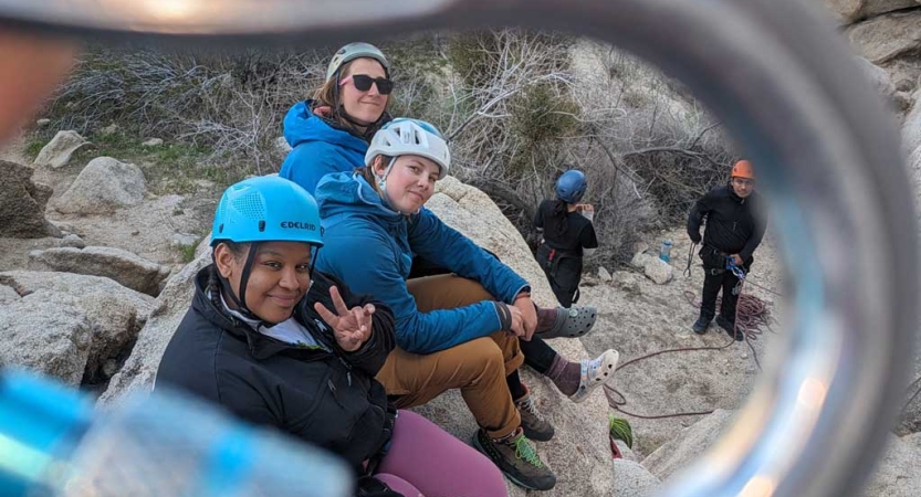 The photo is taken through the center of a carabiner, framing people wearing helmets sitting on a rock and smiling for the photo.    